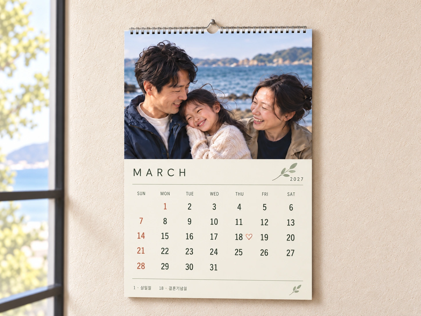 Illustrative wall calendar with a large family photo above an ivory and sage March 2027 layout, terracotta Sundays and Korea’s March 1 holiday, and an anniversary heart beside the 18th