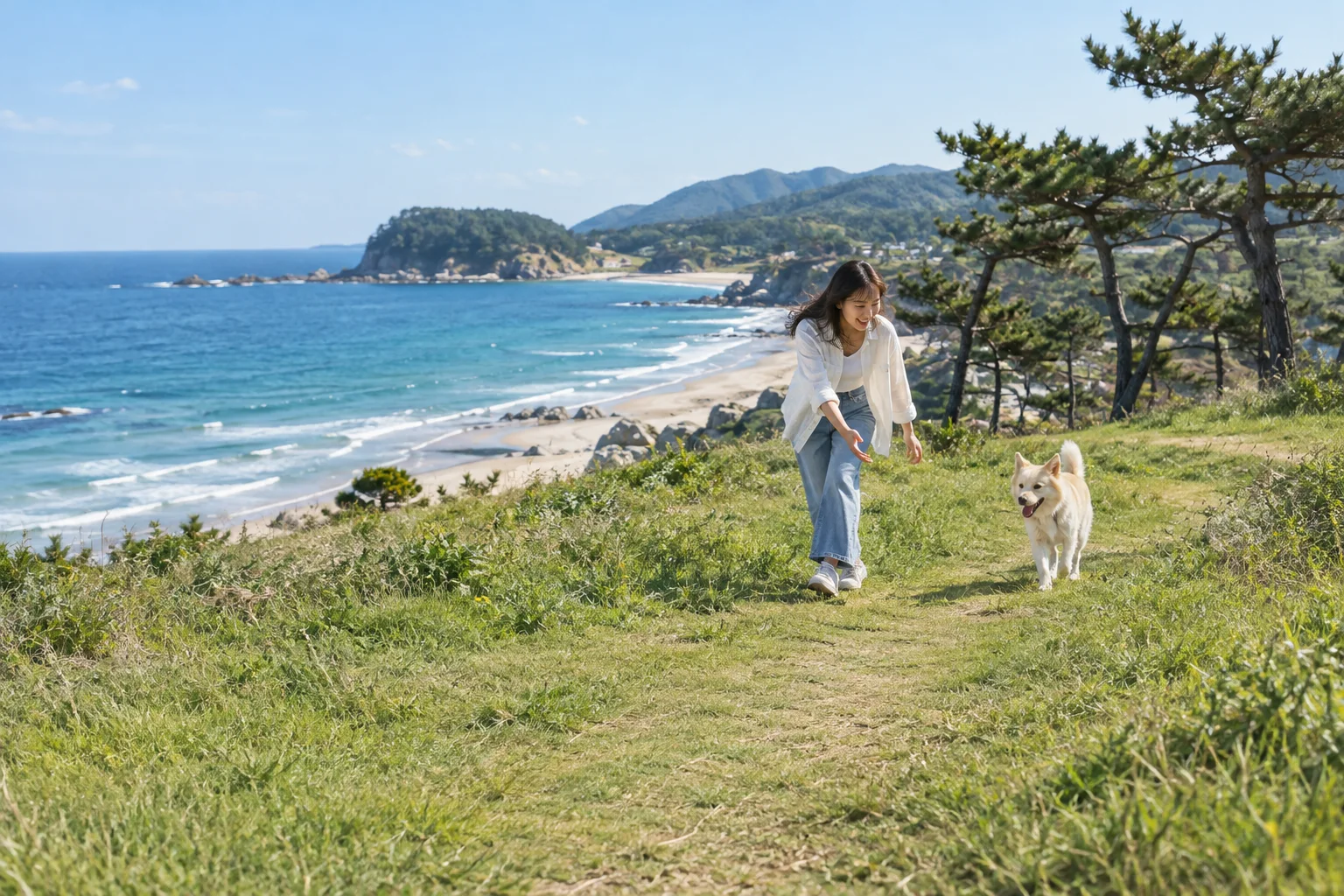 On a grassy headland above the sea on a clear day, a woman leans down to call her dog as it trots toward her